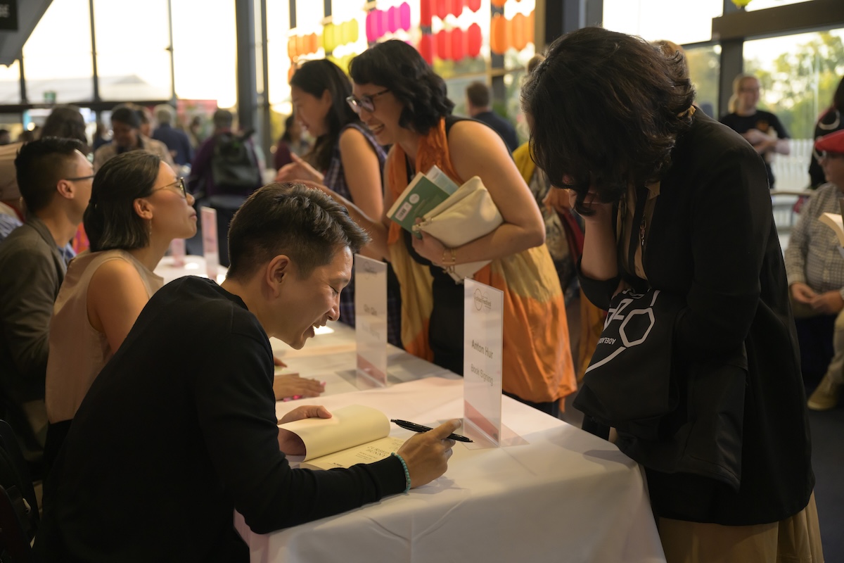 Author signing books for attendees at the OzAsia Festival Weekend of Words event inside a brightly lit venue space.